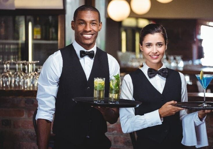 Two people in a restaurant holding trays of drinks.